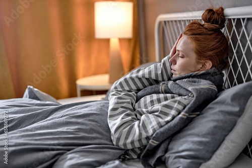 Sick pale tired woman with closed eyes touch forehead, suffering from headache, sitting on bed in sweater, scarf. Young female feeling unhealthy strong sudden pain, morning hangover, closeup side view © alfa27