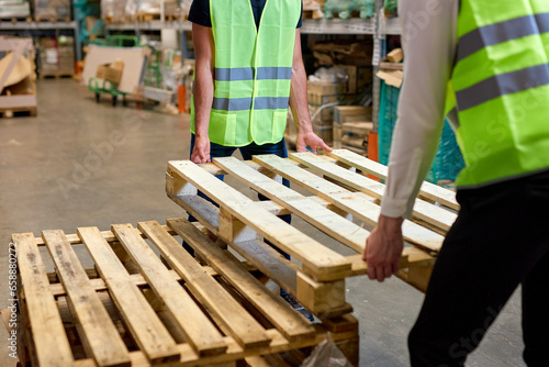 cropped warehouse men hold cargo pallet together with co-worker in workplace area. hardworking strong guys in green vests working uniforms engaged in work in cargo distribution storage