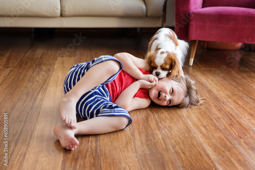 Laughing young boy laying down on wooden floor while his dog is licking his face