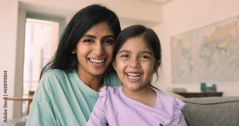 Cheerful beautiful Indian woman cuddling, tickling her cute little daughter, hugging sit together on sofa posing staring at camera, enjoying carefree leisure. Happy motherhood, loving family portrait Cheerful beautiful Indian woman cuddling, tickling her cute little daughter, hugging sit together on sofa posing staring at camera, enjoying carefree leisure. Happy motherhood, loving family portrait