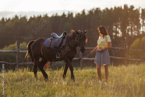 A young girl walks across a field. Portrait of a rider and a black horse against the background of a forest at sunset