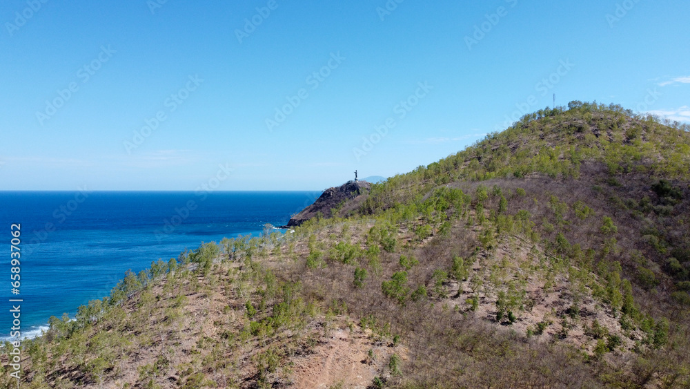 Aerial scenic view of Cristo Rei Jesus Christ statue hiding behind the hill landscape with blue ocean in capital city of Dili, Timor-Leste, Southeast Asia