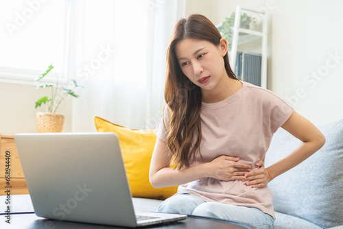 Telehealth, Telemedicine concept. Flatulence asian patient young woman stomach ache suffer from food, discussing with doctor video call with computer at home, during online consultation in living room