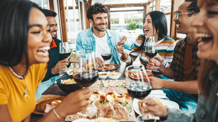 Happy friends drinking red wine sitting at restaurant table - Multiracial young people enjoying rooftop dinner party together - Food and beverage concept with guys and girls having lunch break outside