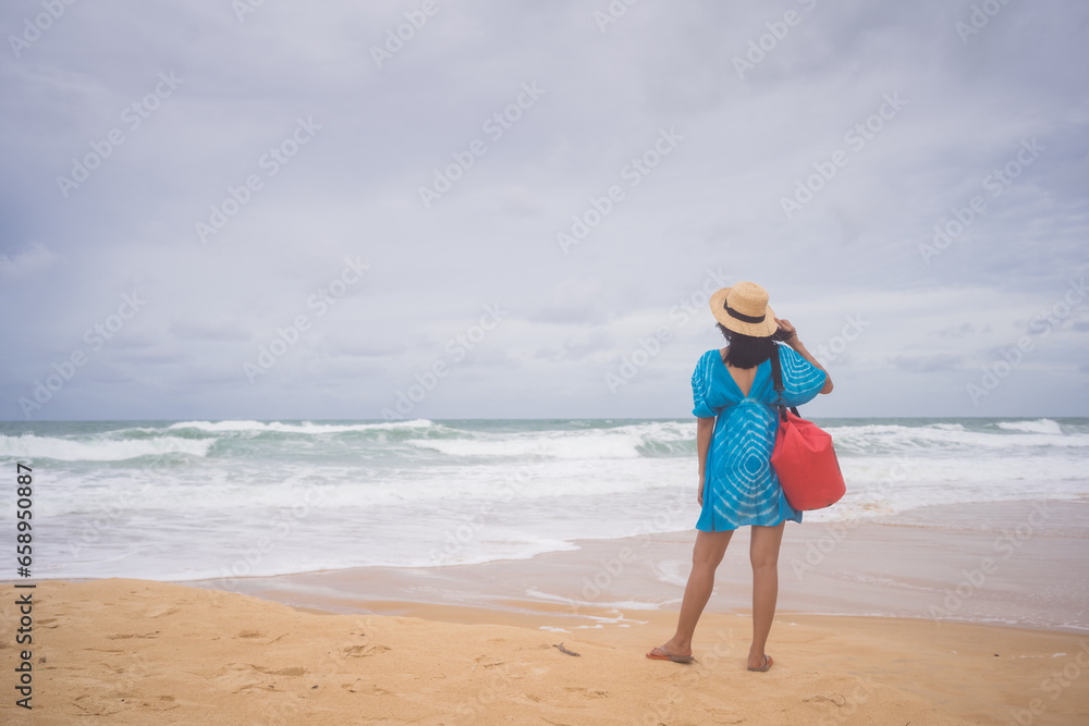 Female tourist in a blue dress carrying a red bag stands on the beach, White waves and blue sky in phuket, Thailand. Female tourist in a blue dress carrying a red bag stands on the beach, White waves and blue sky in phuket, Thailand.