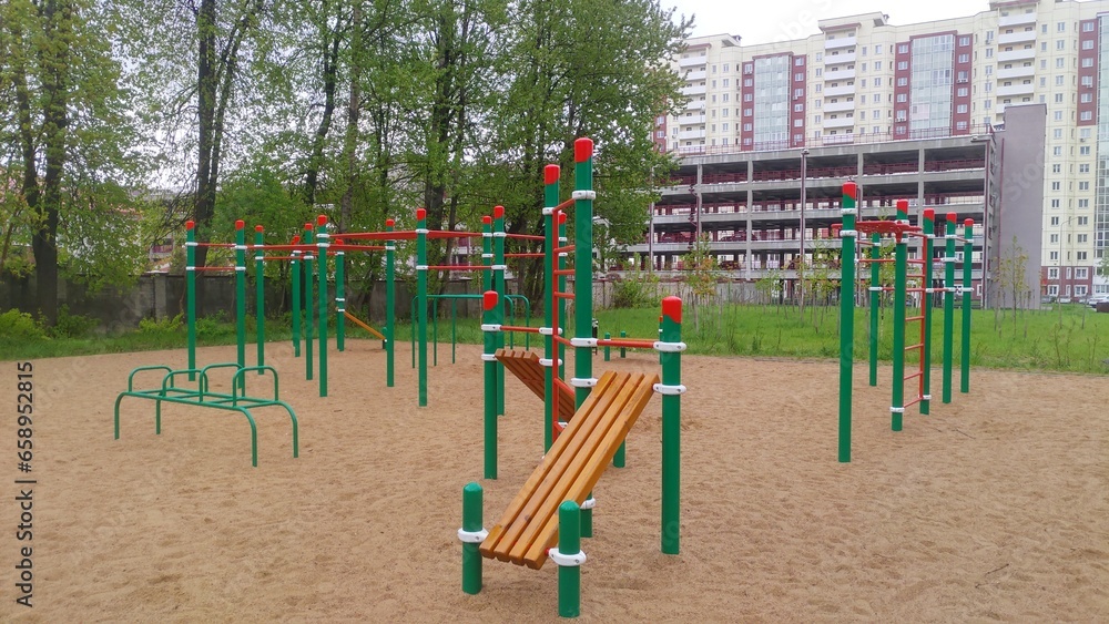 A variety of metal exercise equipment for general use is set up on a sandy area near the lawn, trees, residences and multi-level parking lot. Spring day and cloudy weather A variety of metal exercise equipment for general use is set up on a sandy area near the lawn, trees, residences and multi-level parking lot. Spring day and cloudy weather