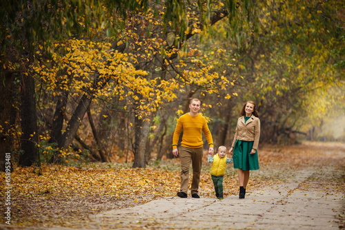 Family dressed up in autumn-style look walks in autumn landscape. Alley covered with yellow foliage. Autumn walk outdoors. Mom, Dad and little son in autumn park. Happy family time. Happy relatives