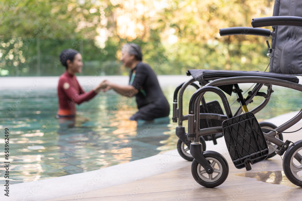 Fototapeta premium Selective focus off wheelchair background of physical therapist helping senior woman in aqua, old women and mature doing aqua aerobics exercise in swimming pool, Elderly sports, and active lifestyle.