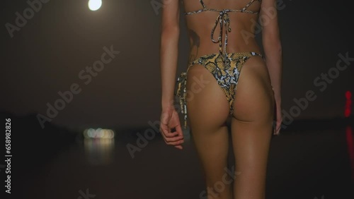 back view of beautiful young adult sexy booty woman in snake print bikini walks in sea at night. fashionable model with perfect slim body in swimsuit at night. summertime adventure.