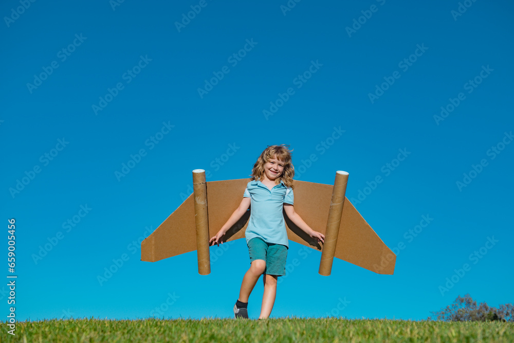 Happy child with paper wings against blue sky. Kid with toy jetpack having fun in spring green field outdoor. Freedom carefree and kids imagination dream concept.