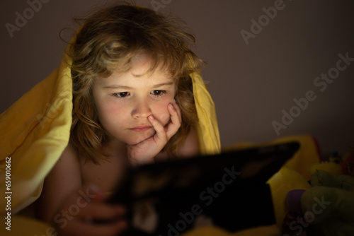 Kid playing game on tablet in bed at night. Kids with social media. Child lying in bed playing a tablet in dark room, light under blacket. Close up of kid watching cartoons on tablet. photo