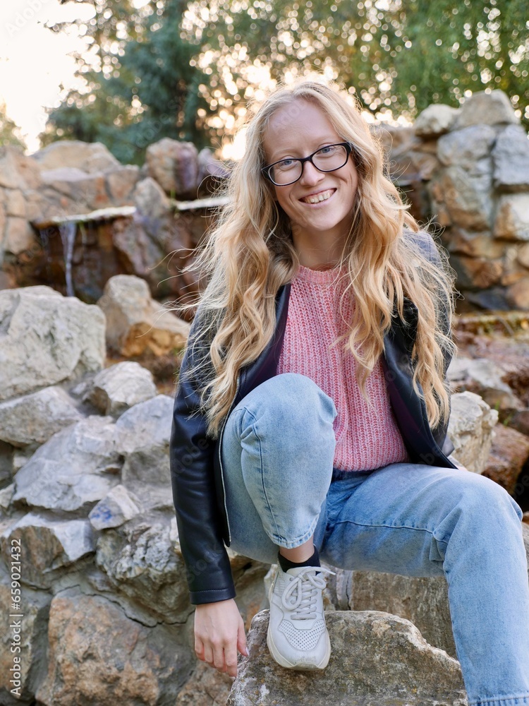 Obraz premium Natural Curly Blonde Woman, Albino Sitting Outdoors and Smiling. Lack of melanin pigment in hair and skin. A person with poor eyesight wearing glasses. Vertical
