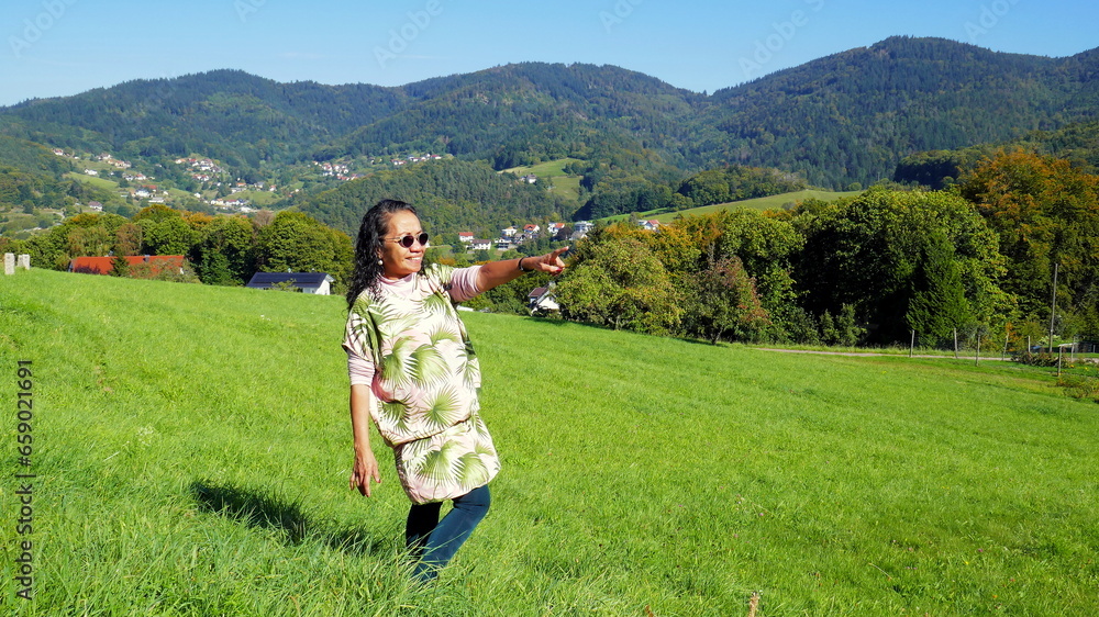 fröhliche Asiatin genießt Aussicht im Schwarzwald bei Sasbachwalden auf Wiese vor bewaldeten Bergen