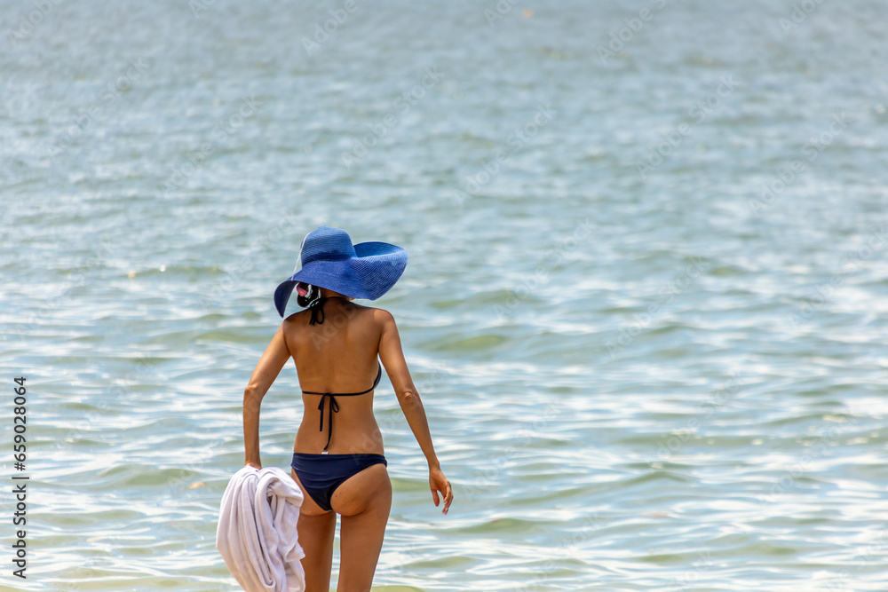 The woman wearing bikini and blue hat standing on the beach of sea to start swimming in Pattaya city in Thailand.