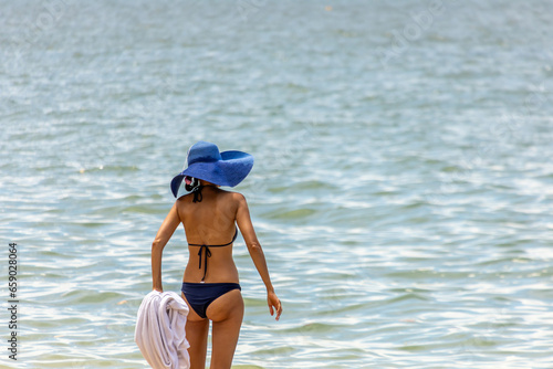 Obraz na płótnie The woman wearing bikini and blue hat standing on the beach of sea to start swimming in Pattaya city in Thailand. w salonie