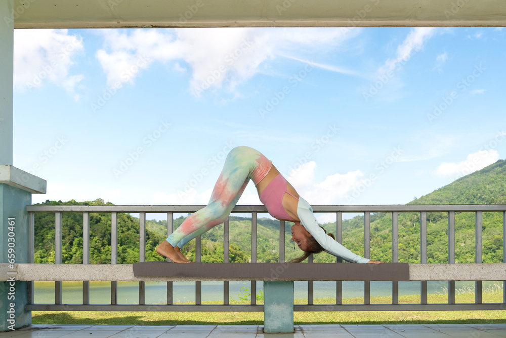 mature blonde woman in vibrant colored yoga suit performs standing stretch on bay shore. Close-up, bright sunlight. Concept of health, fitness, self-care, morning exercise, yoga instructor