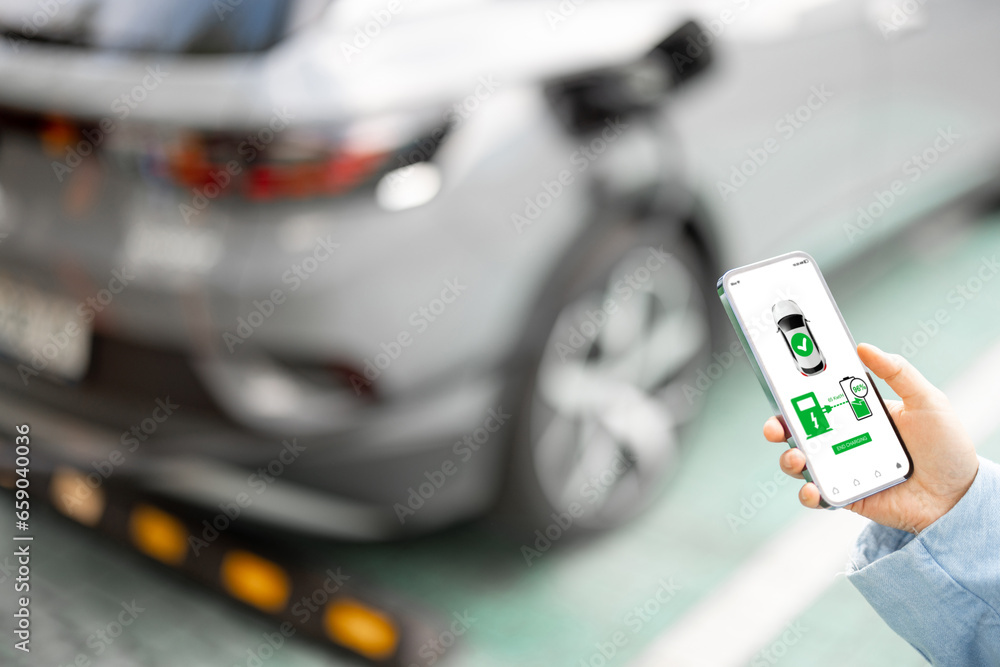 Woman holds a smartphone with running application for vehicle charging, electric car charging on background, close-up