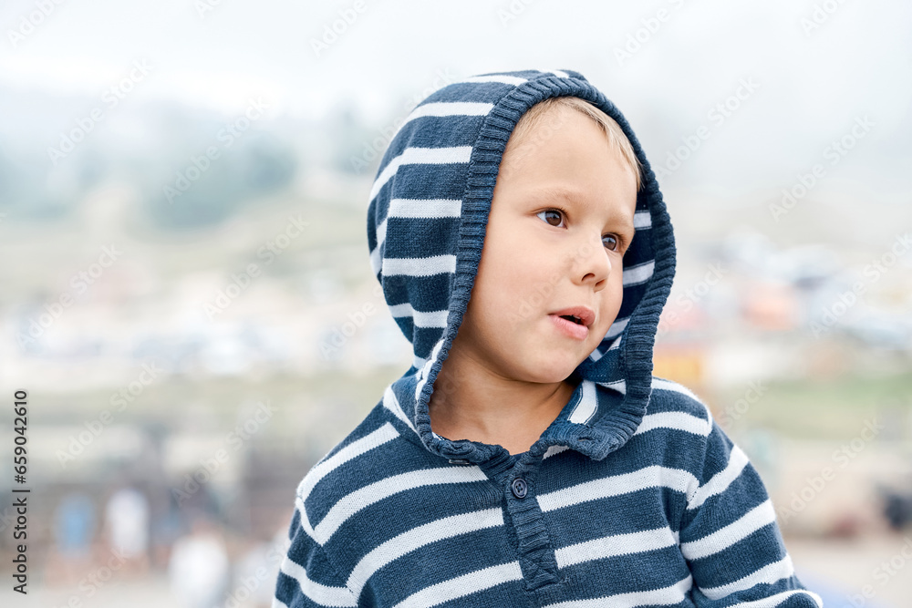 Playful little boy wearing hoodie makes face on blurry background outdoor. Funny child puts head on shoulder walking in mountain park. Happy childhood Playful little boy wearing hoodie makes face on blurry background outdoor. Funny child puts head on shoulder walking in mountain park. Happy childhood