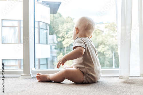 Curious baby looks out of panoramic window sitting on floor. Adorable toddler child rests near large window in hotel room. Healthy kid at home
