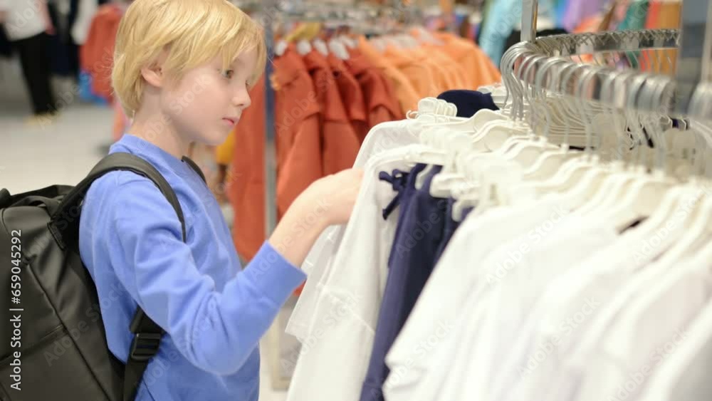 A pretty preteen age boy chooses fashion clothes for himself in a store. Shopping for family with children. An independent child helps his mother in a shopping mall A pretty preteen age boy chooses fashion clothes for himself in a store. Shopping for family with children. An independent child helps his mother in a shopping mall