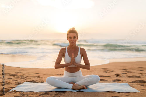 Sunset meditation. Lady meditating outdoors on sea beach, sitting on fitness mat and keeping hands in namaste gesture