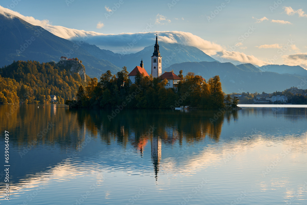 A calm autumn morning on Lake Bled. An island with a church in the foreground and a castle and mountains in the background.