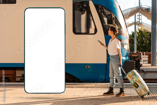 Side view of young woman walking with her luggage and using cellphone. Train station in background. Big smartphone with mock up and copy space. Concept of travel, business trip and vacation