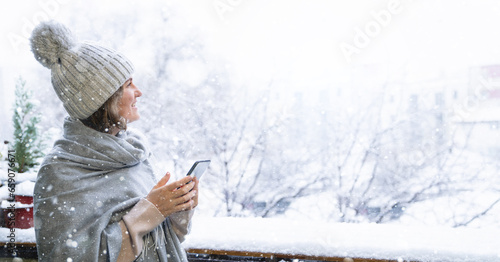 A woman in a warm hat and blanket holds smartphone in her hands. Winter landscape with snowfall in the background