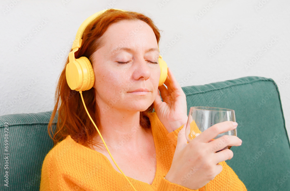Woman with yellow headphones and a glass of wine sitting on the sofa at home and listening to music or podcast. music and relaxation