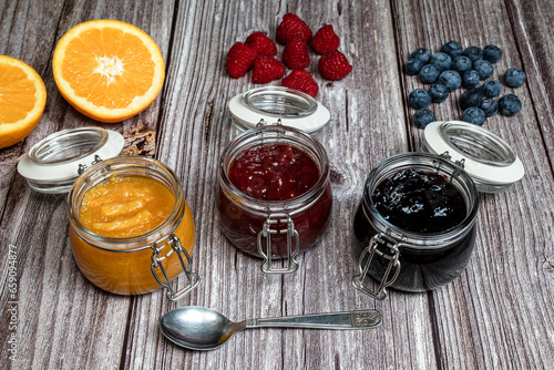 Assortment of homemade jams. Jams in glass jars, oranges, strawberries and blueberries. Front view of seasonal fruits in jam or marmalade, teaspoon.
