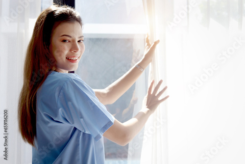 Free Download Side view of happy smiling beautiful woman doctor with stethoscope in blue coat standing and opening white curtain at window in clinic hospital. Young female nurse and medical health care worker. - GFXLoad
