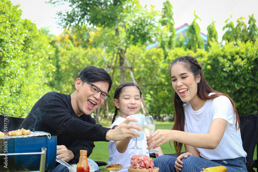 Fototapeta premium Joyful parent and kid child make barbecue at picnic party, have fun together in summer green garden, father mother and daughter girl enjoy eating and drinking at backyard, happy family love bonding.