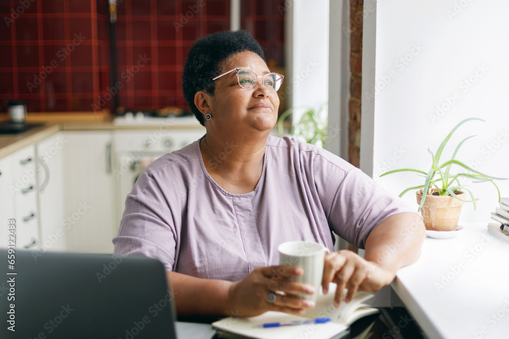 ensive thoughtful senior plus size woman of african ethnicity in spectacles sitting at kitchen table with coffee in hands looking through window contemplating, having rest after working on laptop ensive thoughtful senior plus size woman of african ethnicity in spectacles sitting at kitchen table with coffee in hands looking through window contemplating, having rest after working on laptop
