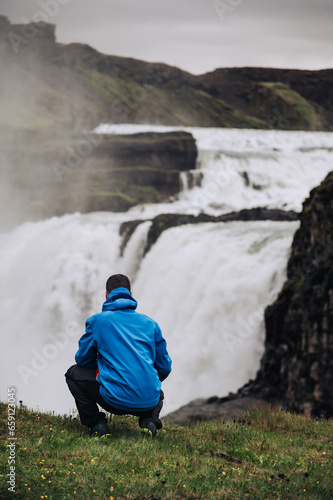 Rear view of a man crouching on a cliff and looking at the waterfall in Iceland. © Zamrznuti tonovi