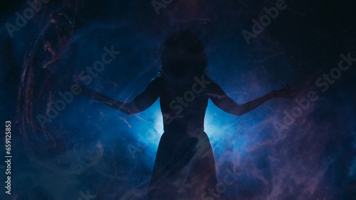 Spotlight silhouette. Spiritual energy. Woman with spread arms in haze shadow blue projector glowing particles fog dark background copy space.