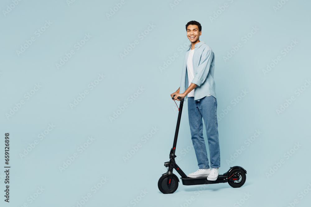 Full body side profile view young man of African American ethnicity wear shirt casual clothes riding electric scooter look camera isolated on plain pastel light blue cyan background studio portrait. Full body side profile view young man of African American ethnicity wear shirt casual clothes riding electric scooter look camera isolated on plain pastel light blue cyan background studio portrait.