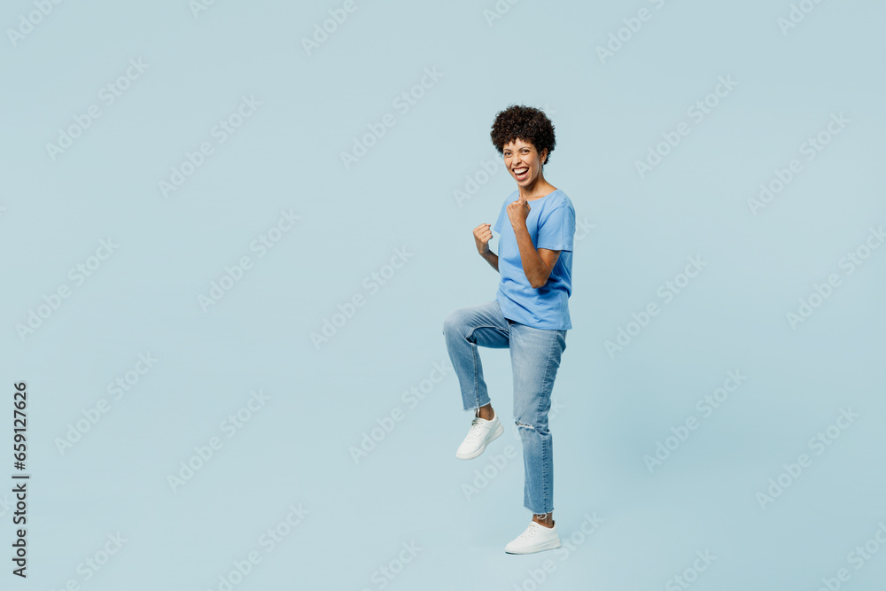 Full body young woman of African American ethnicity wear t-shirt casual clothes doing winner gesture celebrate clenching fists say yes isolated on plain light blue cyan background. Lifestyle concept. Full body young woman of African American ethnicity wear t-shirt casual clothes doing winner gesture celebrate clenching fists say yes isolated on plain light blue cyan background. Lifestyle concept.