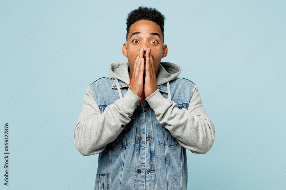 Young surprised man of African American ethnicity wear denim jacket orange t-shirt cover mouth with hand look camera isolated on plain pastel light blue background studio portrait. Lifestyle concept. Young surprised man of African American ethnicity wear denim jacket orange t-shirt cover mouth with hand look camera isolated on plain pastel light blue background studio portrait. Lifestyle concept.
