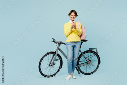 Full body young happy woman student wear casual clothes sweater backpack bag ride bicycle hold in hand use mobile cell phone isolated on plain blue background. High school university college concept.