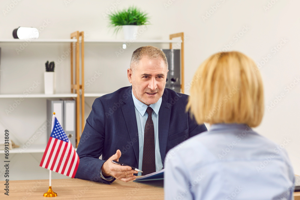 Woman sitting back in the office of the US public services or embassy having consular visa interview with an elderly mature officer with US flag at the desk of official's workplace. - obrazy, fototapety, plakaty 