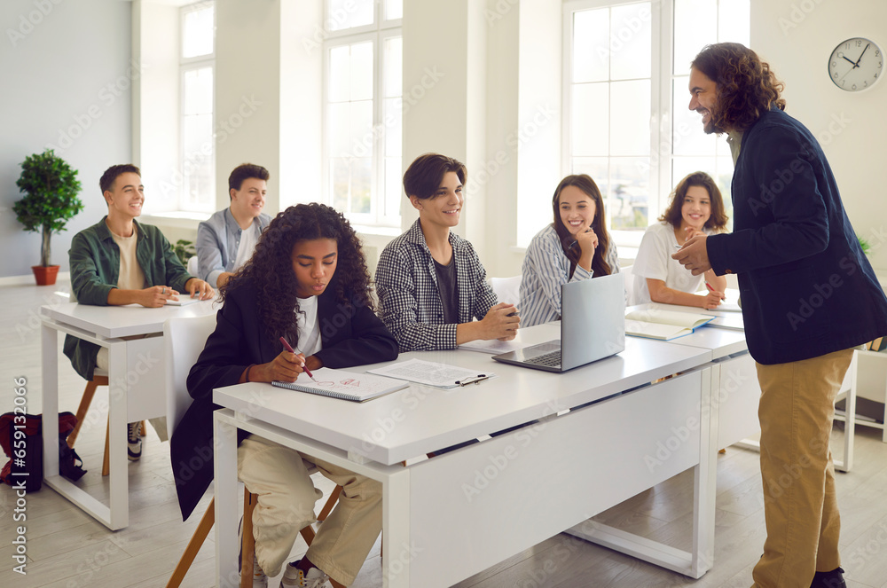 Male teacher conducts lesson for high school students and discusses topic of lesson with them. Smiling group of students sitting at desks in classroom and listening to teacher. Education concept. Male teacher conducts lesson for high school students and discusses topic of lesson with them. Smiling group of students sitting at desks in classroom and listening to teacher. Education concept.