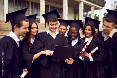 Portrait of a happy diverse multiethnic graduate students in black graduation robes celebrate graduation and looking to diploma of their classmate and laughIng in university campus outdoors.