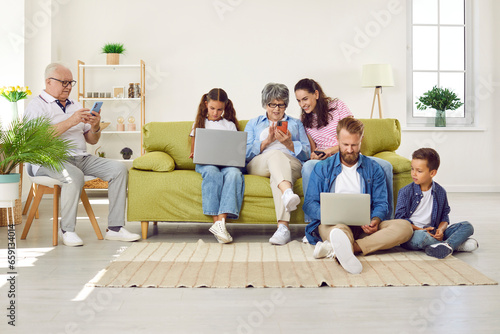 Big family sitting in living room with gadgets, parents, grandparents and two children. Grandpa, mom and grandma looking at smartphones screens, daughter and father working on laptops with son.