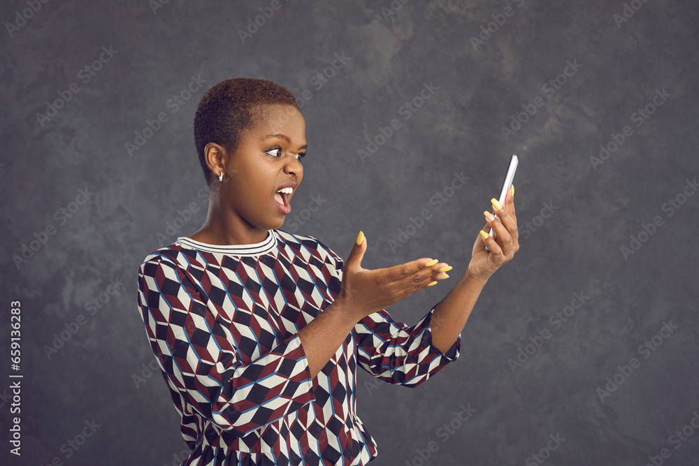 Portrait of dissatisfied and embarrassed dark-skinned woman who has problems with her mobile phone. Woman standing on gray background outraged by virus attack, lack of mobile communication or internet Portrait of dissatisfied and embarrassed dark-skinned woman who has problems with her mobile phone. Woman standing on gray background outraged by virus attack, lack of mobile communication or internet