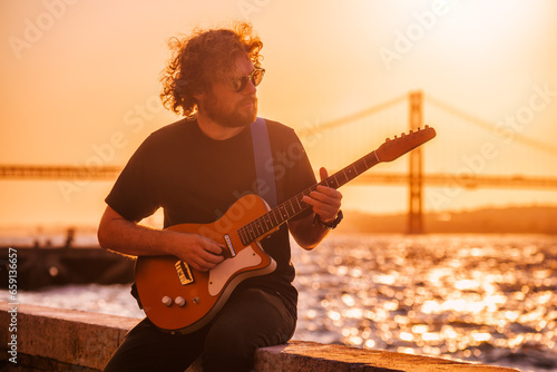 Print op canvas Hipster street musician in black playing electric guitar in the street on sunset on embankment with 25th of April bridge in background