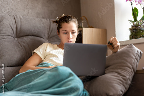 Attractive woman sitting comfy sofa under blanket at home and browsing laptop.