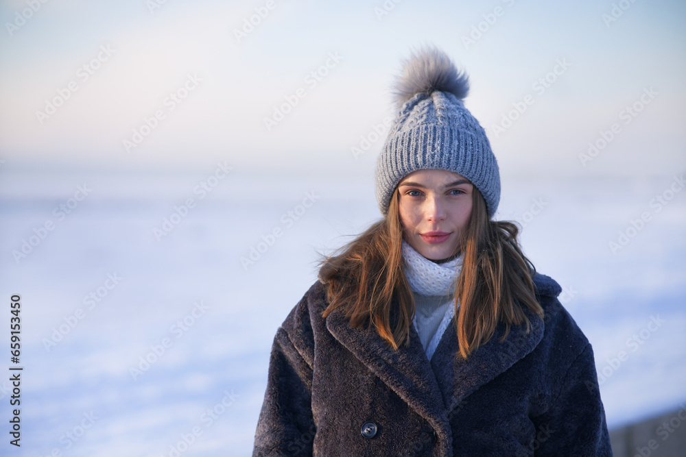 Portrait of beautiful girl young frozen pretty woman standing walking in winter snowy park at cold snow frosty day in hat, scarf, in gloves looking at camera Portrait of beautiful girl young frozen pretty woman standing walking in winter snowy park at cold snow frosty day in hat, scarf, in gloves looking at camera
