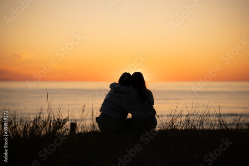 Silhouette of two female friends enjoying a conversation while sitting hugging and watching the sunset on a beach. Back view photo