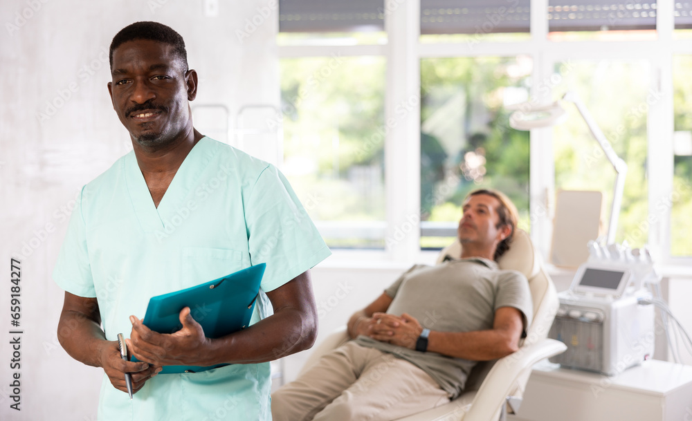 Fototapeta premium Middle-aged African man doctor stands with arms crossed on chest in hospital office, medical center. Modern medical care, tests, consultation