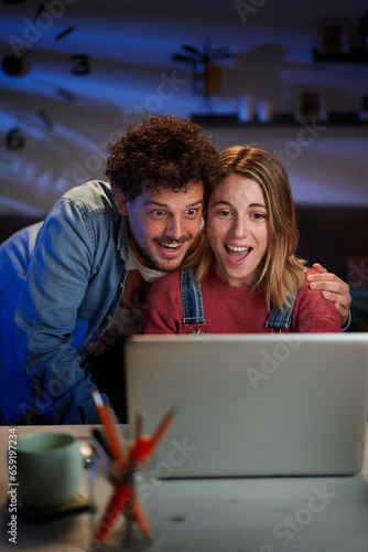Vertical surprised Caucasian couple looking happy at computer sitting at desk living room at night. Excited millennial people having fun at home using laptop. Positive relationships, leisure moments.